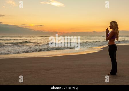 Giovane donna bionda in forma da sola praticando yoga su una spiaggia all'alba o al tramonto Foto Stock