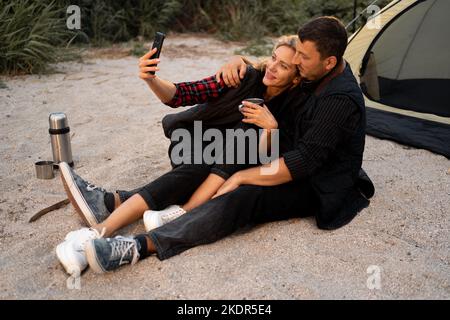 Coppia di campeggio in una tenda che prende un selfie. Coppia felice che fa selfie mentre si siede al campeggio. Persone, stile di vita e tecnologia Foto Stock