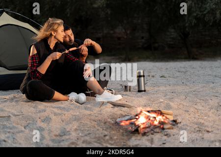 Giovane coppia cucinando salsicce alla legna, facendo un pic-nic al campeggio sulla spiaggia la sera. Felice coppia e falò in riva al mare Foto Stock