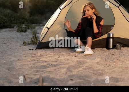 Donna felice in tenda mangiare salsiccia fritta sul fuoco mentre riposa nel campo. Fare un picnic al campeggio sulla spiaggia in serata. Campeggio, cibo Foto Stock