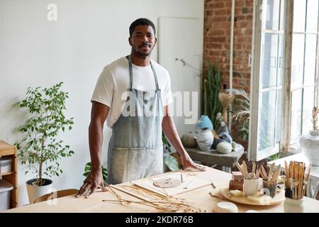 Vita in su ritratto di giovane uomo nero che indossa grembiule mentre si trova in ceramica studio e sorridendo alla macchina fotografica, copia spazio Foto Stock