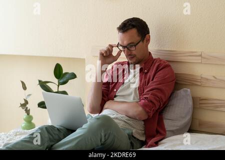 L'uomo caucasico concentrato si siede sul letto nella camera dell'hotel leggendo le notizie via internet o controllando la posta elettronica Foto Stock