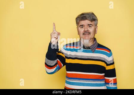 Uomo maturo con barba grigia accigliata e punta il dito verso l'alto isolato su sfondo giallo Foto Stock
