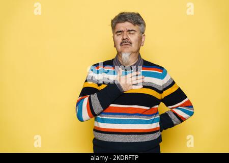 Uomo triste maturo con barba grigia che tiene la mano sul petto isolato su sfondo giallo Foto Stock