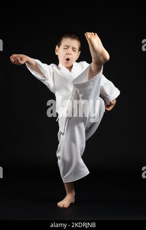 Un ragazzino in un kimono pratica il karate su sfondo nero, calciando in avanti. Foto Stock