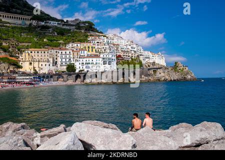 La città di Amalfi, sulla costiera amalfitana, si affaccia dal molo di pietra che protegge il porto con 2 nuotatori che guardano. Foto Stock