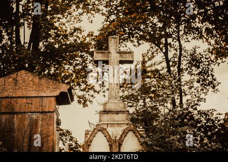 Un'immagine scura di una vecchia croce di pietra su una lapide in un cimitero sullo sfondo di alberi e cielo nuvoloso. Religione. Foto Stock