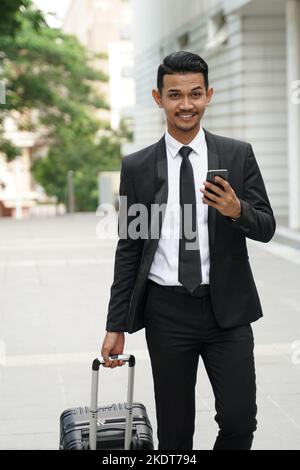 Elegante uomo d'affari che controlla la posta elettronica sul telefono cellulare mentre cammina con la valigia Foto Stock