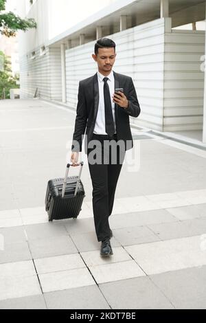 Elegante uomo d'affari che controlla la posta elettronica sul telefono cellulare mentre cammina con la valigia Foto Stock