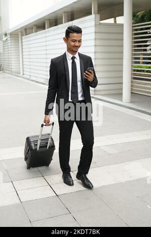 Elegante uomo d'affari che controlla la posta elettronica sul telefono cellulare mentre cammina con la valigia Foto Stock