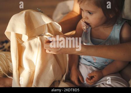 Bambina che succhia un succhietto seduto sul pavimento e guarda la madre ricamatrice. Tra generazioni. Decorazione domestica. Primo piano ritratto. Foto Stock