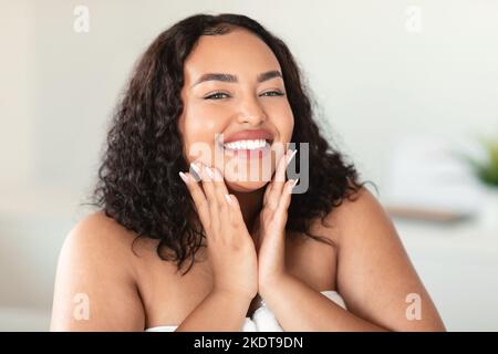 Ritratto di bella donna nubby nera che tocca le guance e sorride alla macchina fotografica, godendo di trattamenti per la cura della pelle a casa Foto Stock