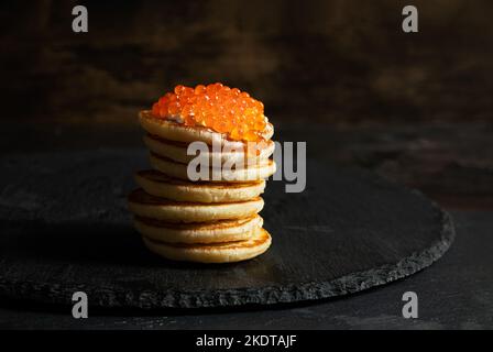 Caviale rosso su piccoli pancake blinis con panna acida su sfondo scuro Foto Stock