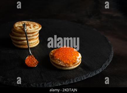 Caviale rosso in un cucchiaio e su piccoli pancake blinis con panna acida su ardesia Foto Stock