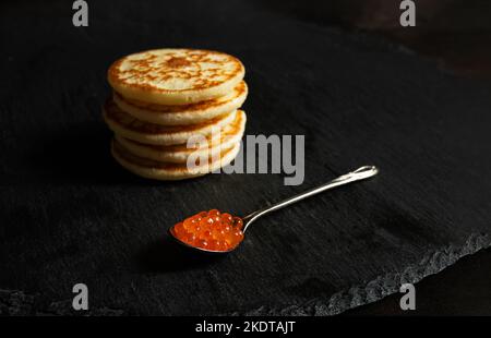 Un cucchiaio di caviale rosso e piccoli pancake blinis su un ardesia Foto Stock