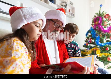 Primo piano di un uomo anziano vestito come Babbo Natale che legge una storia a due bambini a casa. Natale, vacanza. Foto Stock