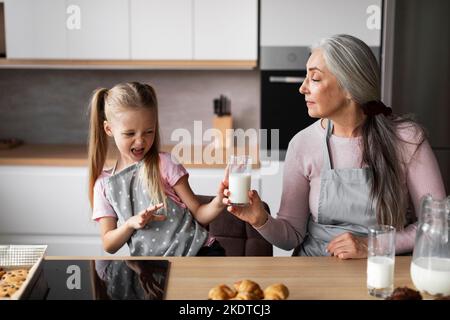 La piccola grimaces caucasica insoddisfatta rifiuta un bicchiere di latte dalla vecchia nonna negli interni moderni della cucina Foto Stock