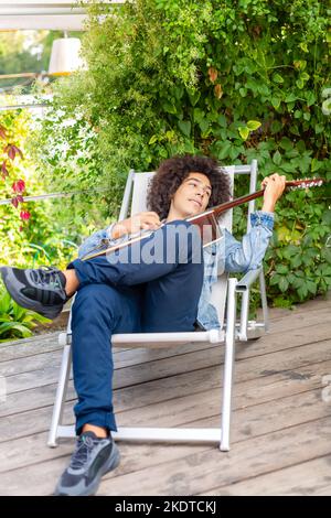 Bel giovane afro-italiano uomo di corsa mista in abbigliamento casual suonare la chitarra in giardino. Un ragazzo multirazziale suona uno strumento musicale riposa e. Foto Stock