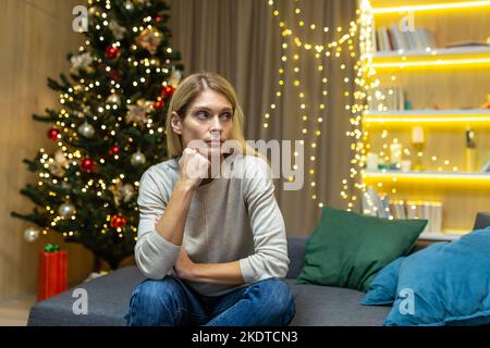 Triste e solitaria giovane bella donna seduta a casa sul divano vicino all'albero di Natale. Spende le feste di Natale da solo. Foto Stock