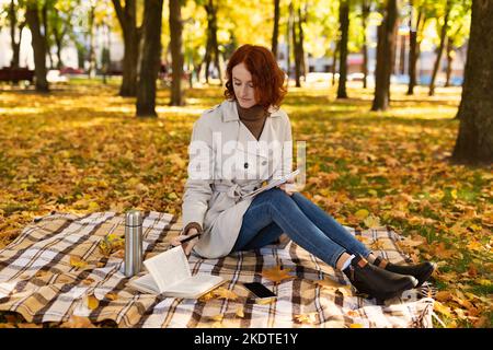 Concentrato intelligente caucasico piuttosto millenario signora studente con capelli rossi in impermeabile siede su plaid, legge libro Foto Stock