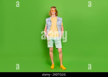 Vista a tutta lunghezza delle dimensioni del corpo del ragazzo allegro attraente hippie tenendo le mani in tasche isolato su sfondo di colore verde brillante Foto Stock