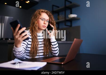 Donna bionda riccia in occhiali che lavora da casa con più dispositivi elettronici Internet. Freelancer donna d'affari tiene tablet, parlando sul cellulare Foto Stock