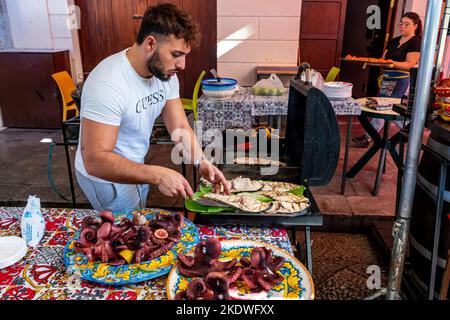 Un giovane uomo cucinare il pesce su Una griglia al Ballaro Street Market, Palermo, Sicilia, Italia. Foto Stock