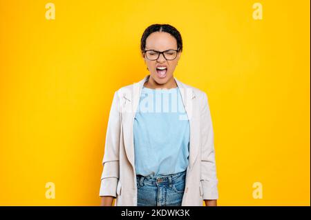 Irritata arrabbiata ispanica o brasiliana, dipendente aziendale, in elegante abbigliamento, con gli occhiali, urla con gli occhi chiusi, in non se stessa dalla rabbia, si trova su sfondo isolato arancione Foto Stock