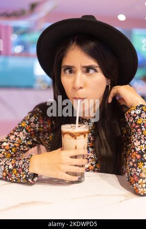 fare un grin e bere una bevanda fredda con una paglia, espressione facciale e bevande, giovane donna latina con capelli lunghi e cappello, stile di vita Foto Stock