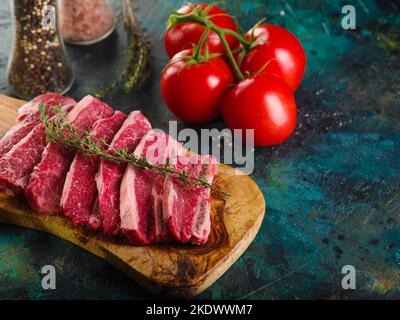 Bistecche di carne con un rametto di rosmarino profumato su un tagliere di legno, spezie, condimenti in vasetti di vetro e pomodori maturi su sfondo marmoreo. Re Foto Stock