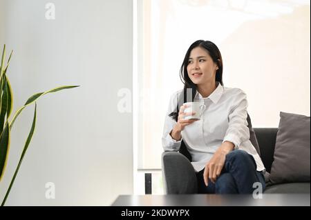 Attraente giovane donna asiatica rilassa sorseggiando caffè del mattino mentre si rilassa nel suo salotto. Foto Stock