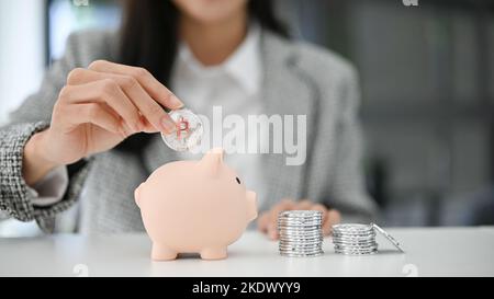 Immagine ravvicinata, bella donna d'affari asiatica inserire le monete in un pin piggy bank sul tavolo. Risparmio di denaro, investimenti, concetto di valuta Foto Stock