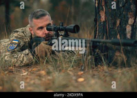 Soldato ucraino vestito uniforme militare sdraiato a terra con fucile da cecchino con silenziatore in prima linea. Bell'uomo coraggioso e fiducioso in guerra Foto Stock