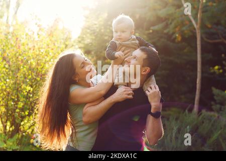 Madre, papà e bambino figlio che trascorrono il tempo insieme nel parco estivo godendosi il caldo sole. Il bambino si siede sulle spalle dei padri. Giovane caucasico f Foto Stock