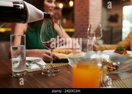 Cameriere che versa vino rosso in bicchiere per una donna seduta al tavolo da pranzo Foto Stock