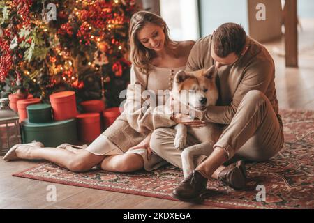 Ritratto di giovane bella coppia abbracciando la vigilia indoor 25 dicembre con il cane akita inu. La gente in maglia elegante hugs vestito bacia tenerezza celebrando ne Foto Stock