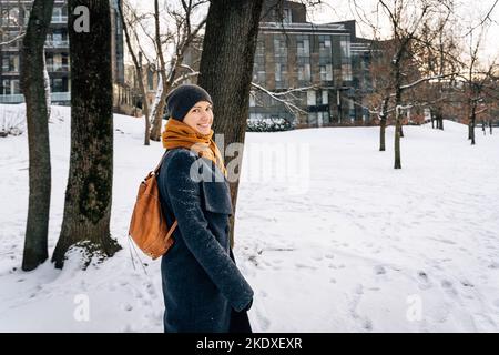 Una donna in abiti invernali con uno zaino in pelle rossa guarda intorno sorridente. Foto Stock
