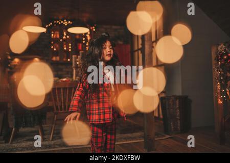 Attiva ragazza bambino giappone con elegante modello di capelli brunette lunga posa a casa in bokeh ghirlands noel. Asian piccolo fashionista in rosso plaid pajama danza Foto Stock