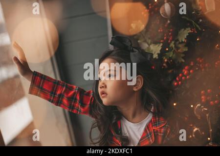 Felice giappone piccola ragazza sognatore guardando attraverso la finestra e miracolo in attesa da Santa posa vicino a noel albero. Bambino asiatico in abito rosso plaid festeggiamento Foto Stock