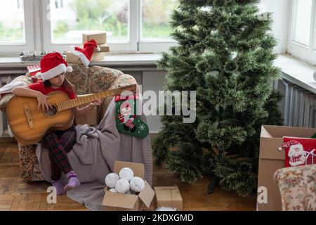 Ragazza piccola felice in cappello rosso giocando il sullo sfondo dell'albero di natale. Il bambino di talento che si diverte durante le vacanze invernali in casa accogliente decorato. Foto Stock