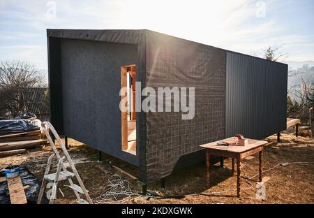 Casa con struttura in legno su fondamenta in pile nel casale in costruzione in stile scandinavo. Parete con membrana impermeabile e lamiera di ferro corrugata nera utilizzata come facciata del futuro cottage. Foto Stock