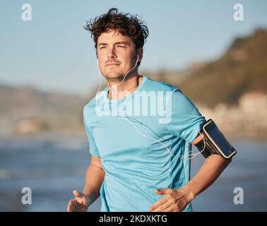 Corsa, musica e atleta uomo in spiaggia con cuffie gestite dall'oceano con streaming audio. Radio mobile, podcast Web o brano di allenamento di un runner Foto Stock