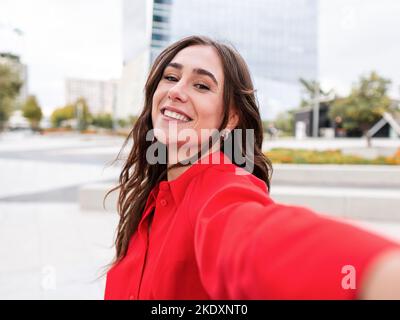 Bruna giovane ottimista in elegante camicia rossa con lunghi capelli castani guardando la fotocamera con sorriso mentre si prende selfie in città strada Foto Stock