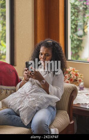 Ragazza indiana che naviga sul cellulare mentre si siede sul divano in un soggiorno luminoso a casa Foto Stock