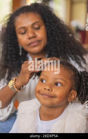 La madre indiana preoccupante con i capelli neri ricci che fanno i ponytail al bambino carino mentre si siede insieme nella stanza luminosa a casa Foto Stock
