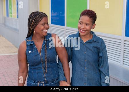 mamma colombiana matura e figlia adulta in abiti denim sorridendo e tenendo le mani mentre camminano vicino all'edificio con pareti colorate sulla strada cittadina Foto Stock