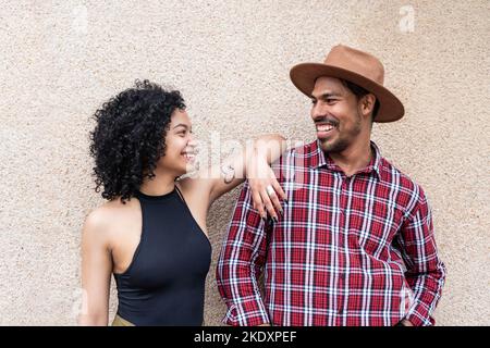 Allegra donna latino-americana in abiti casual con capelli ricci appoggiati sulla spalla del ragazzo e guardarsi con il sorriso mentre si è in piedi vicino Foto Stock