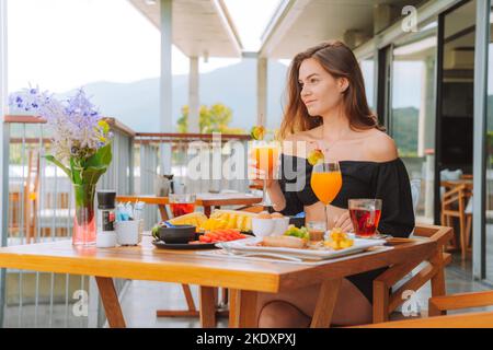 Giovane donna rilassata con un bicchiere di succo d'arancia fresco e una sana e deliziosa colazione con frutta mentre si riposa al tavolo sulla terrazza dell'hotel resort Foto Stock