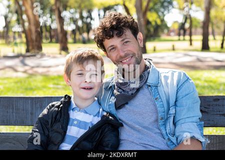 Allegro papà e figlio in abiti casual sorridendo e guardando la macchina fotografica mentre si siede sulla panca su sfondo sfocato del parco illuminato dal sole Foto Stock