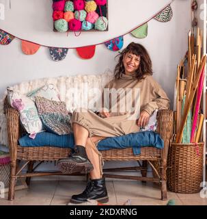 Donna adulta positiva in abito caldo seduta sul divano in vimini in laboratorio con decorazioni appese Foto Stock